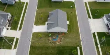 Aerial view of a new suburban house with a worker inspecting a complex network of colored utility lines including water, gas, and fiber optics, highlighting the intricate setup of essential services.